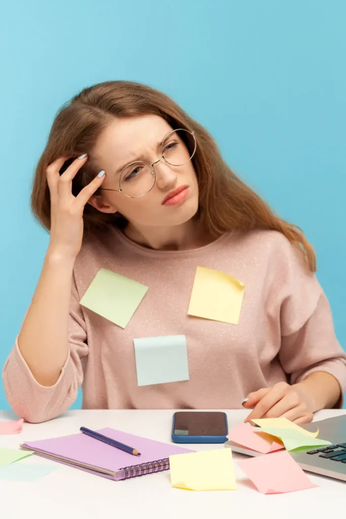 A confused young woman with sticky notes on her clothes, sitting at a desk and looking puzzled, symbolizing memory or focus difficulties.
